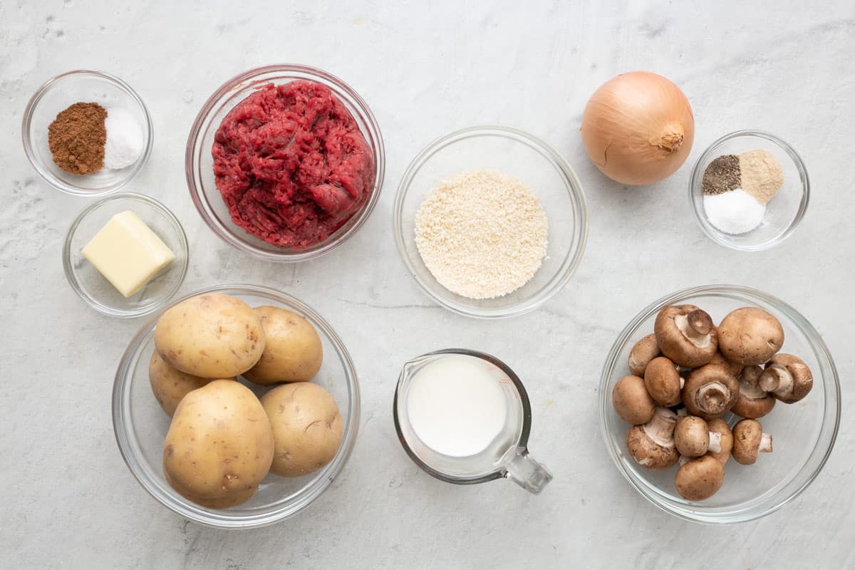Ingredients for recipe in individual bowls: 7 spice and salt, butter, potatoes, ground beef, breadcrumbs, milk, onion, seasonings, and button mushrooms.