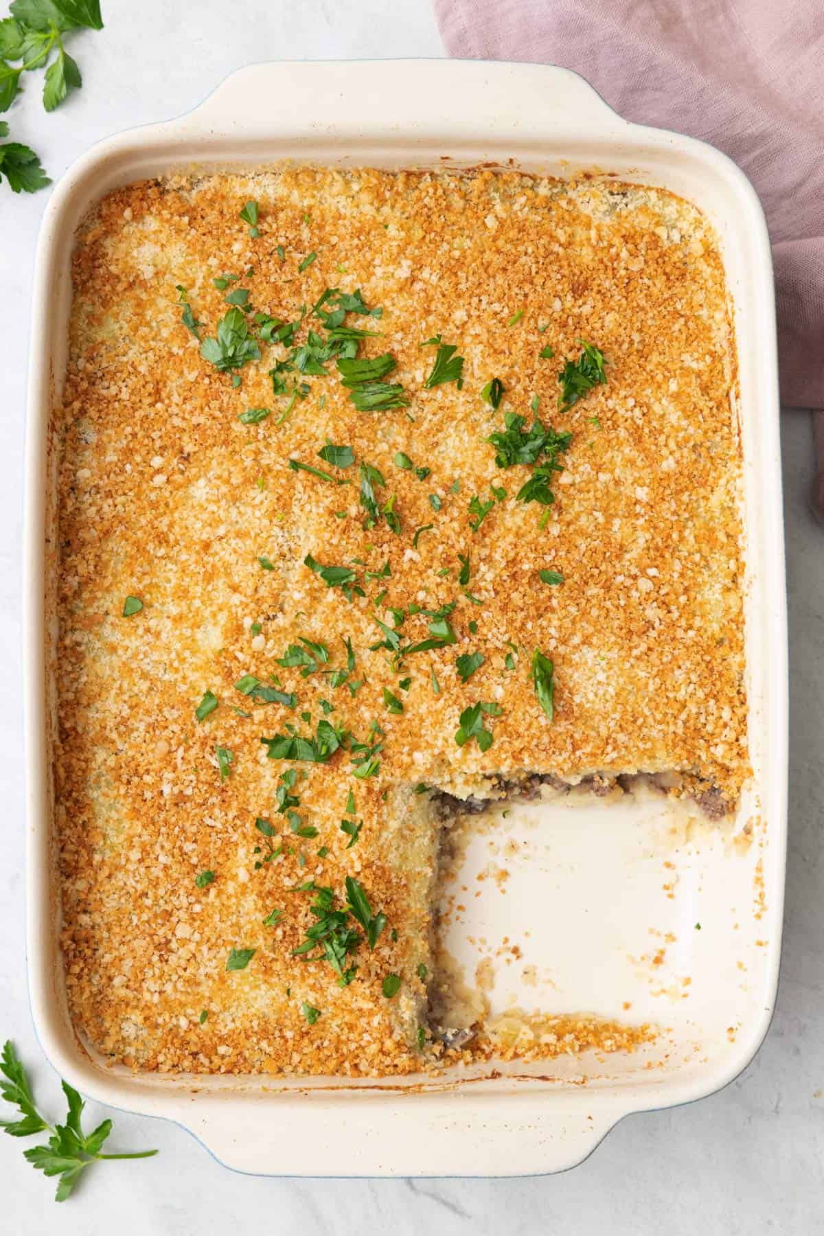 Potato and beef bake in a baking dish with a square serving removed, golden brown breadcrumb, and garnished with fresh chopped parsley.