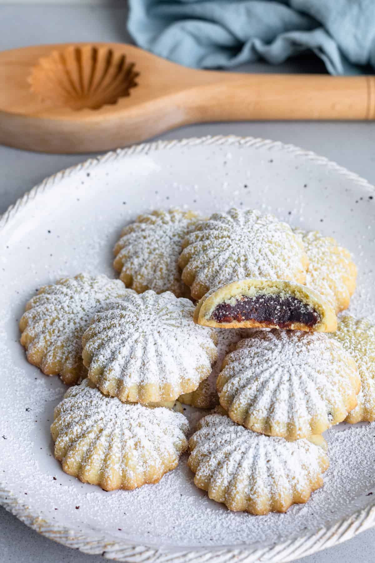 Date paste filled maamoul cookies on a plate with one split open to show inside, dusted with powdered sugar.