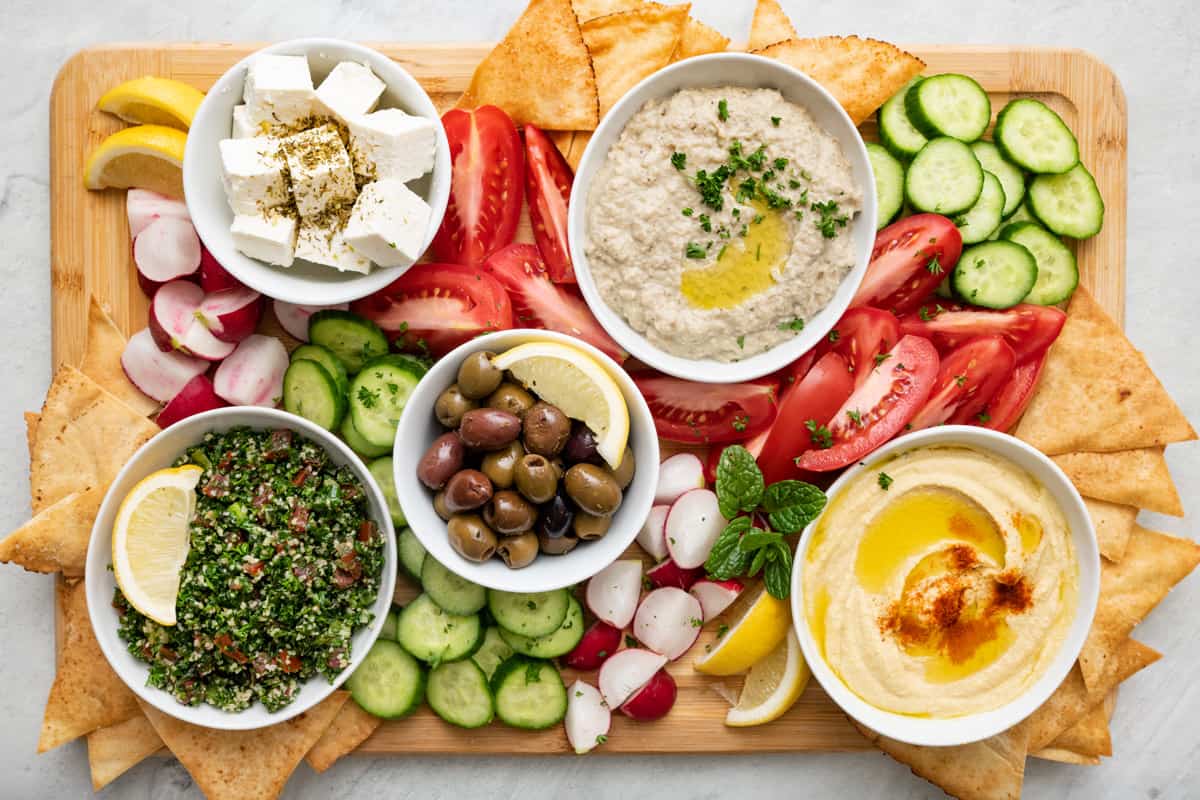 Final mezze platter with small bowls of hummus, baba ghanoush, small blocks of feta, olives, and tabbouleh, with quartered tomatoes and radishes, and sliced cucumbers spread in between bowls. Pita triangles tucked into the edges of the board and lemon garnished throughout.
