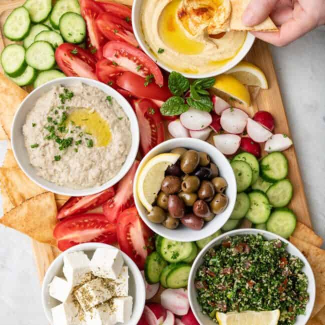 Hand dipping a pita chip into a bowl of hummus from a mezze platter spread with varies dips and vegetables.