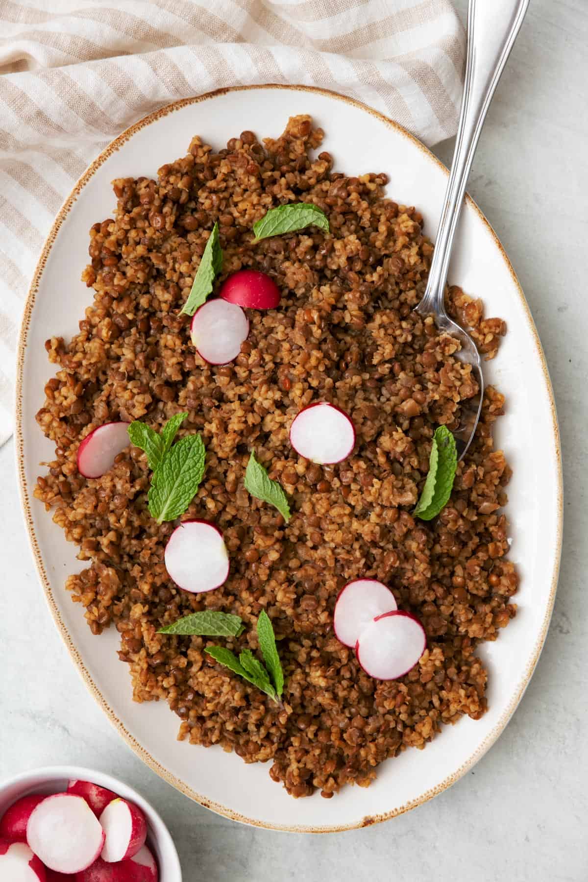 Mujadara hamra in a large shallow serving platter with a serving spoon garnished with fresh mint leaves and a small bowl or radishes cut in half nearby.