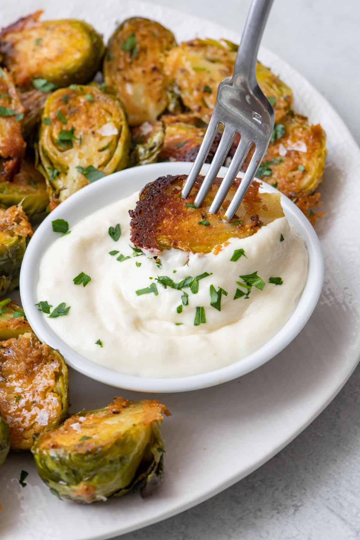 Fork dipping roasted parmesan brussel sprouts into aoili on a plate full of more of them