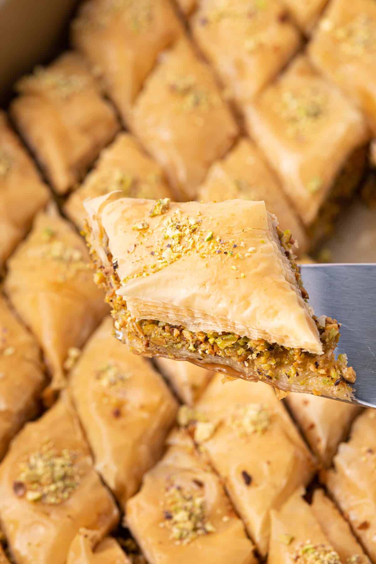 Spatula lifting up a diamond shaped piece of pistachio baklava with full try below it.