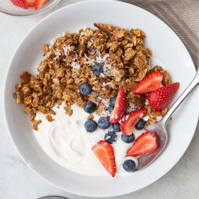 Bowl with scrambled oats, greek yogurt, a few quartered strawberries, blueberries, with coconut sprinkled on top. Small dishes of berries nearby.