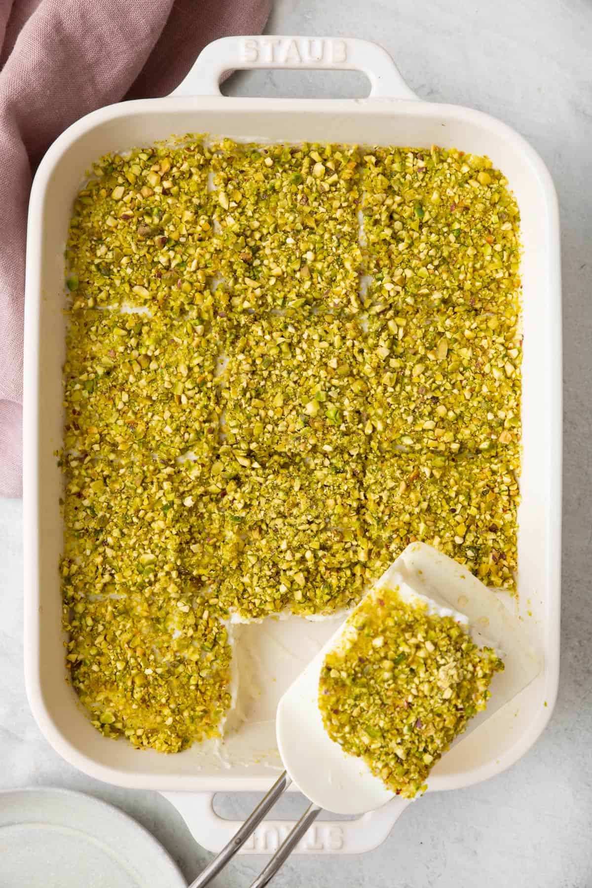 Semolina Pudding topped with pistachios in a rectangular baking dish cut into 12 squares with a spatula lifting a piece out.