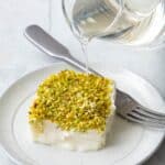 A piece of pudding with chopped pistachios on a plate with simple syrup being poured over from a small clear pitcher.