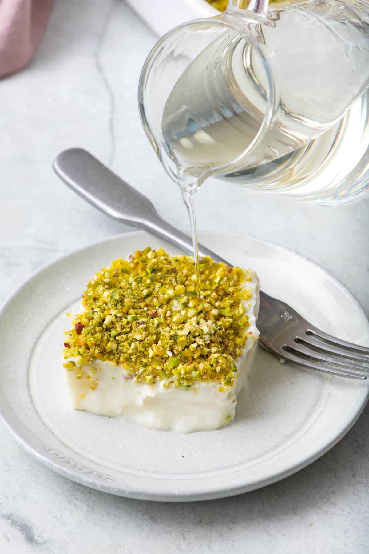A piece of pudding with chopped pistachios on a plate with simple syrup being poured over from a small clear pitcher.