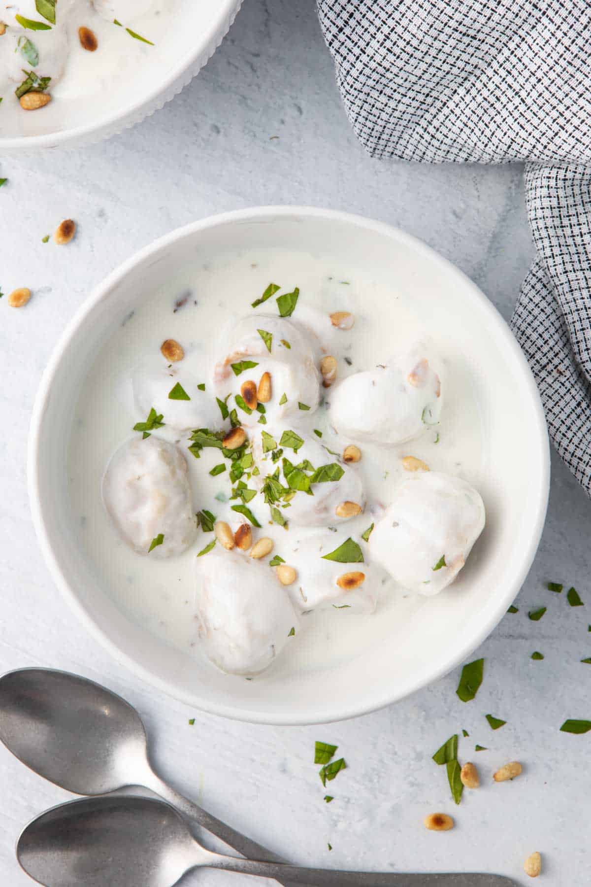 Bowl of shish barak in a creamy yogurt sauce garnished with extra pine nuts and chopped parsley, two spoons resting nearby.