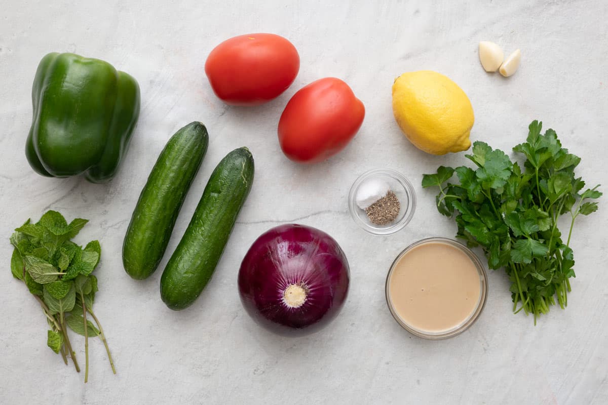 Ingredients for recipe before prepping: fresh mint, green bell pepper, cucumbers, tomatoes, red onion, salt and pepper, lemon tahini paste, garlic, and fresh parsley.
