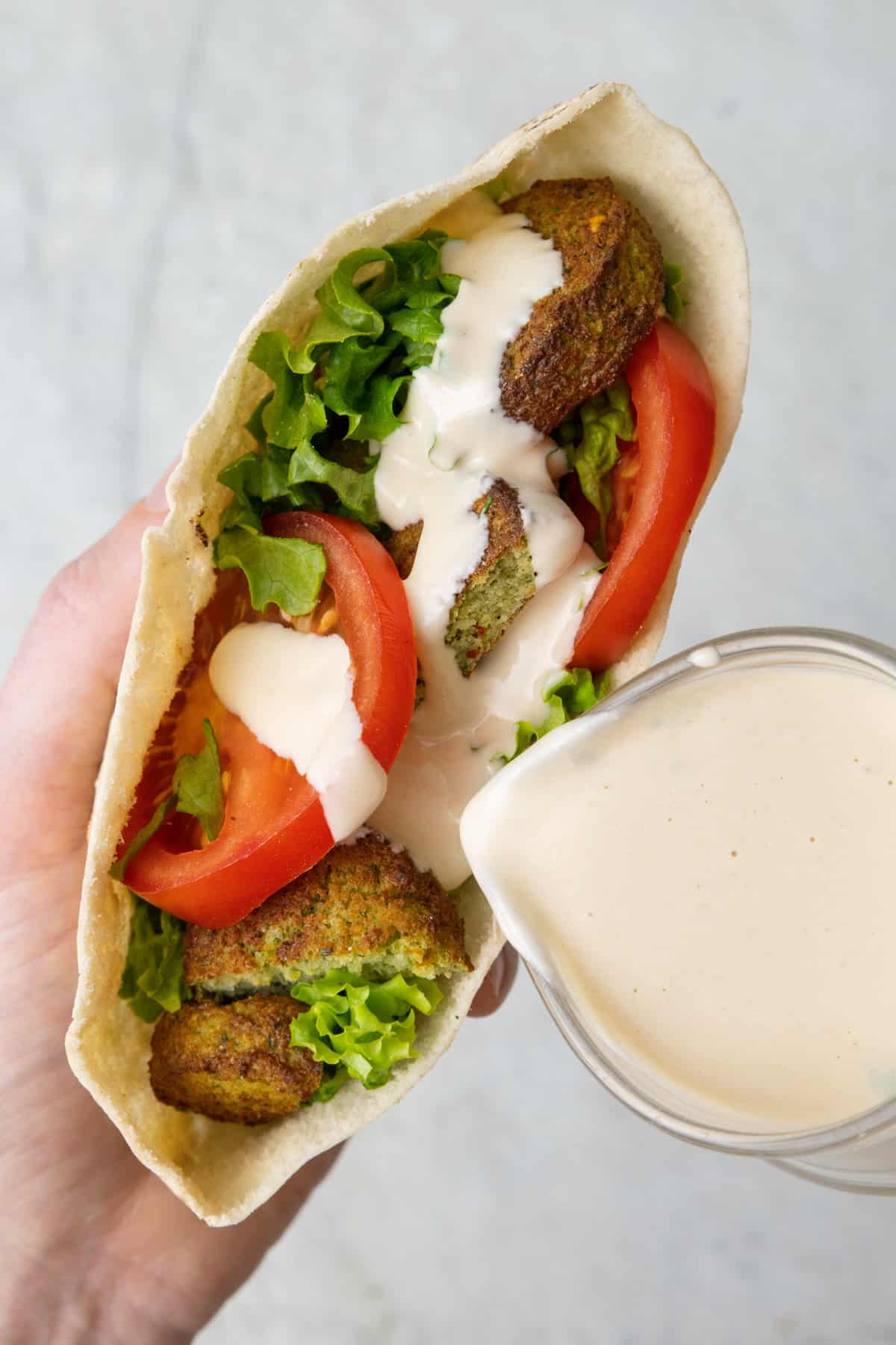 Tahini sauce being poured over a falafel pita stuffed with sliced tomatoes and lettuce.