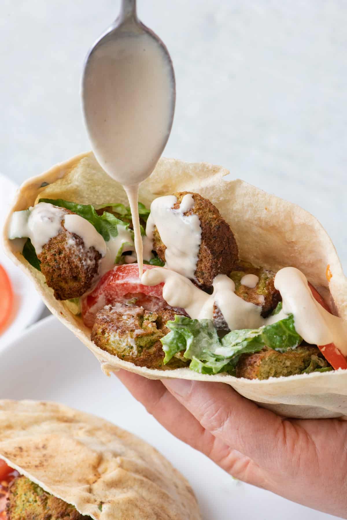 Spoon drizziling tahini over a pita sandwich with falafel, lettuce, and tomato.