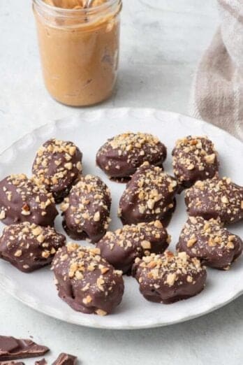 Date Snickers on a round white plate topped with chopped peanuts on top, a small jar of creamy peanut butter with a spoon dipped in and a few chocolate pieces nearby.