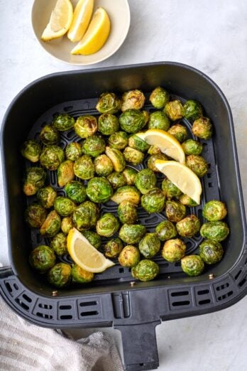 Air fryer Brussel sprouts in an air fryer basket garnished with lemon wedges.
