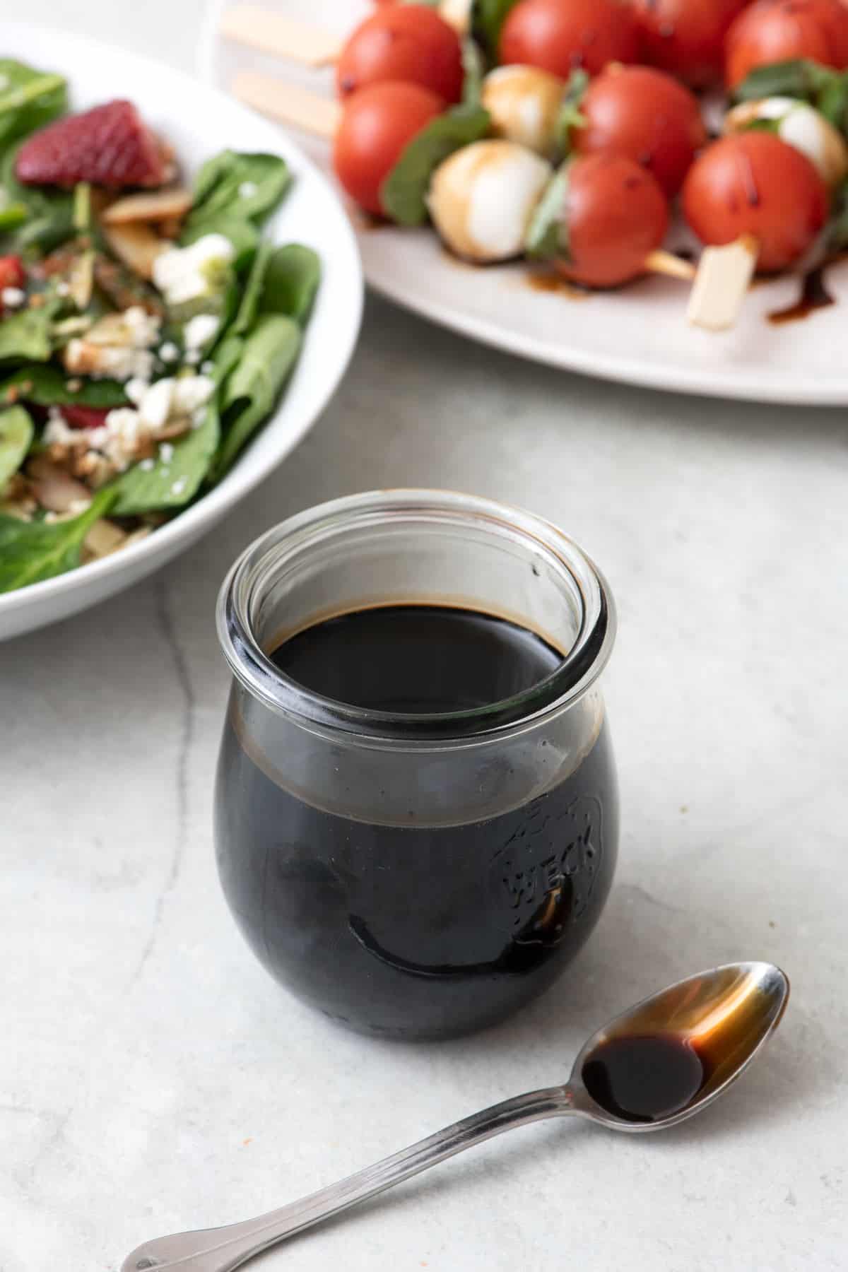 Small jar of balsamic glaze with a spoon sitting nearby. A fresh salad and caprese skewers in the background with the balsamic glaze drizzled on them.