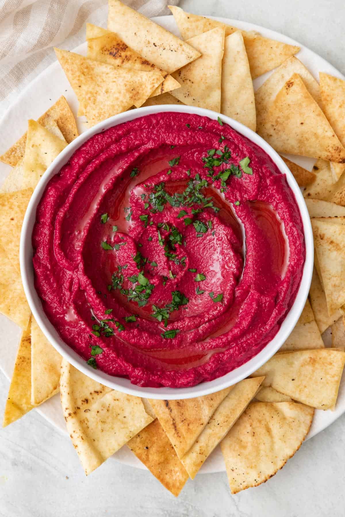 Beet hummus in a bowl garnished with fresh parsley that is set on a plate and surrounded with pita chips.