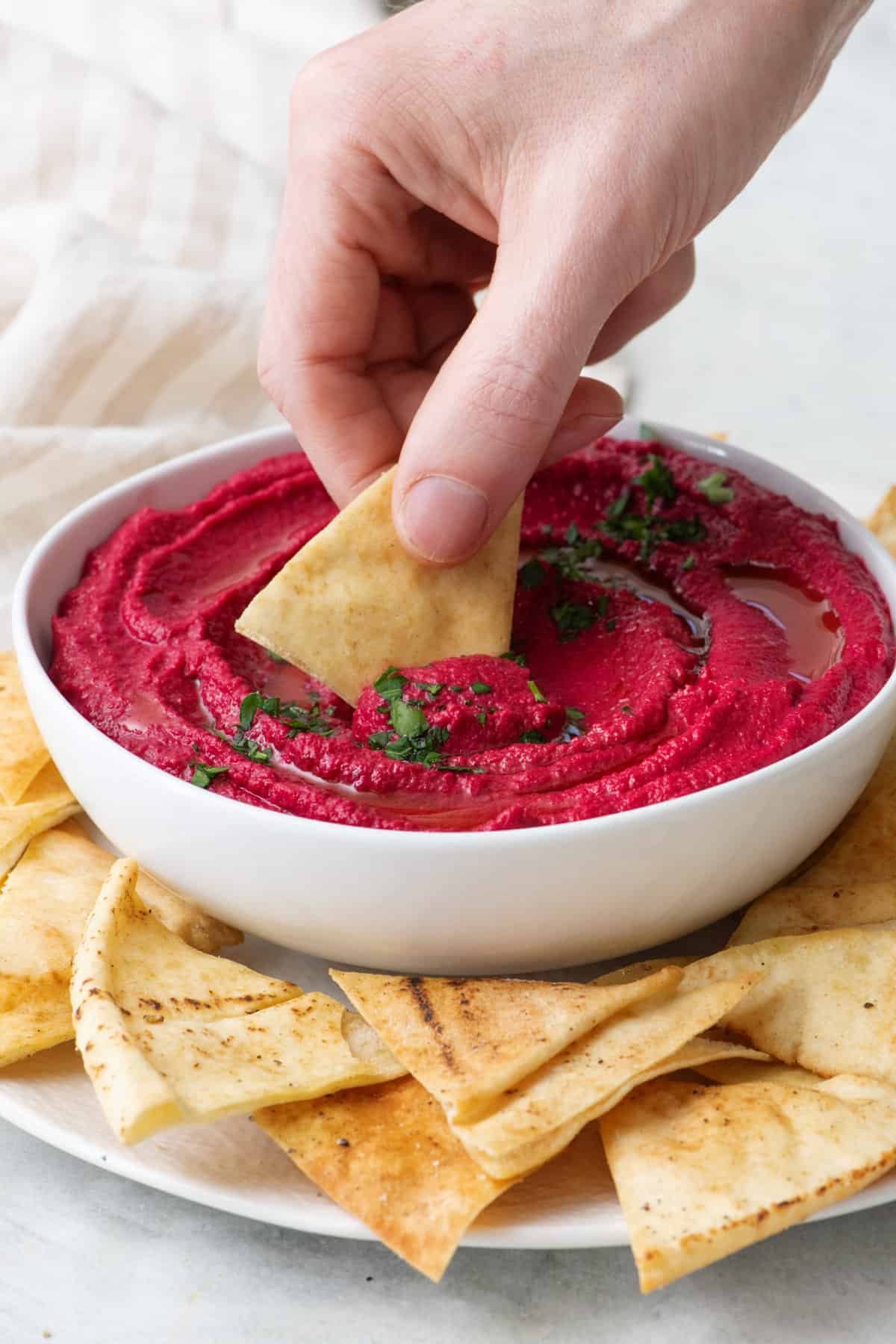 Pita chip being dipped into a dark pink bowl of beet hummus garnished with fresh chopped parsley with more pita chips around.