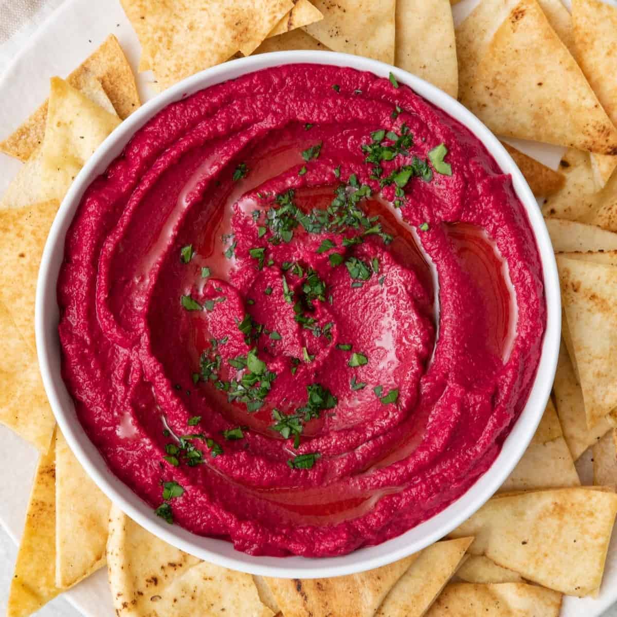 Beet hummus in a bowl garnished with fresh parsley that is set on a plate and surrounded with pita chips.