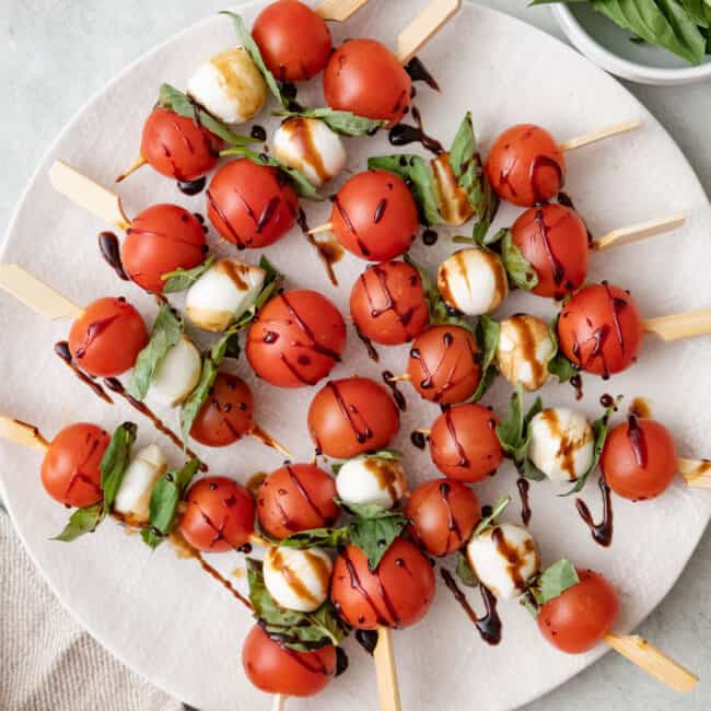 Caprese skewers on a large round plate with balsamic glazed drizzled over and a small dish of additional basil leaves and mozzarella balls nearby.