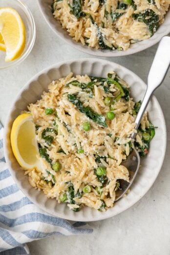 Two small bowls of lemon orzo with peas and spinach garnished with parmesan and lemon wedges.