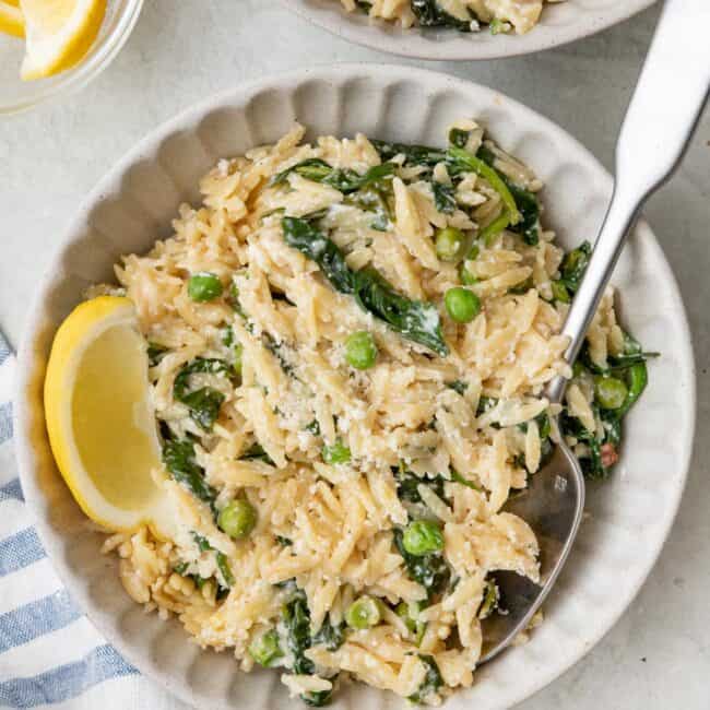 Two small bowls of lemon orzo with peas and spinach garnished with parmesan and lemon wedges.