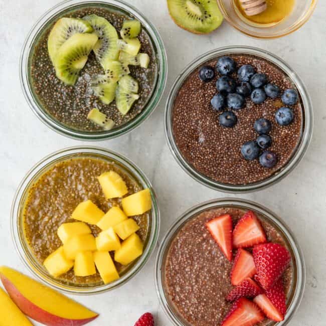 4 different chia jello in bowls: kiwi, blueberry, mango, and strawberry. Extra fruit and a small bowl of honey nearby.