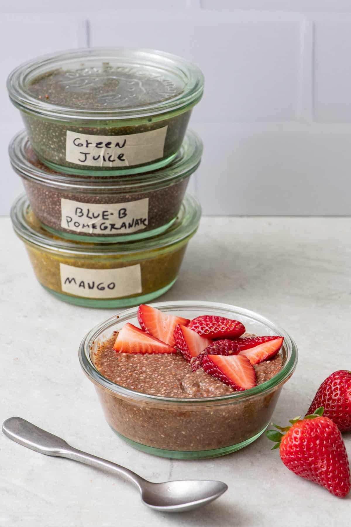 Stack of small glass bowls with lids filled with different chia jello flavors and an unopened chia jello in front topped with strawberries.