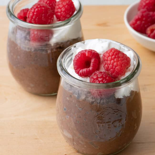 Close up of two glass jars of chocolate pudding topped with whipped coconut cream and raspberries.