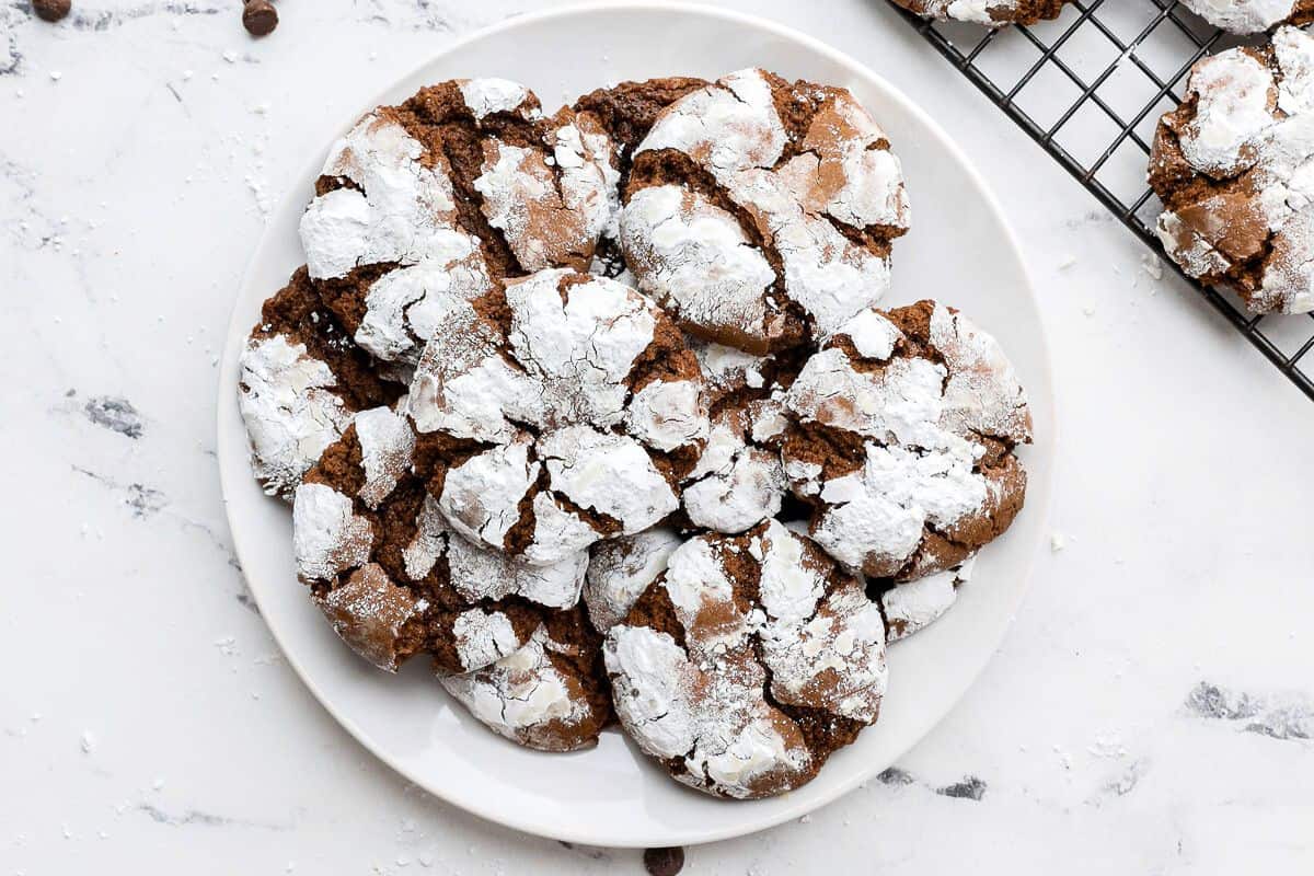 Chocolate crinkle cookies on a round plate with a wire rack of more cookies nearby.
