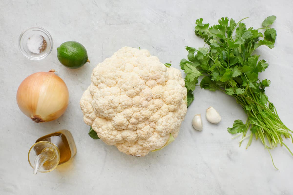 Ingredients for recipe before prepping: salt and pepper, yellow onion, lime, oil, head of cauliflower, garlic, and fresh cilantro.