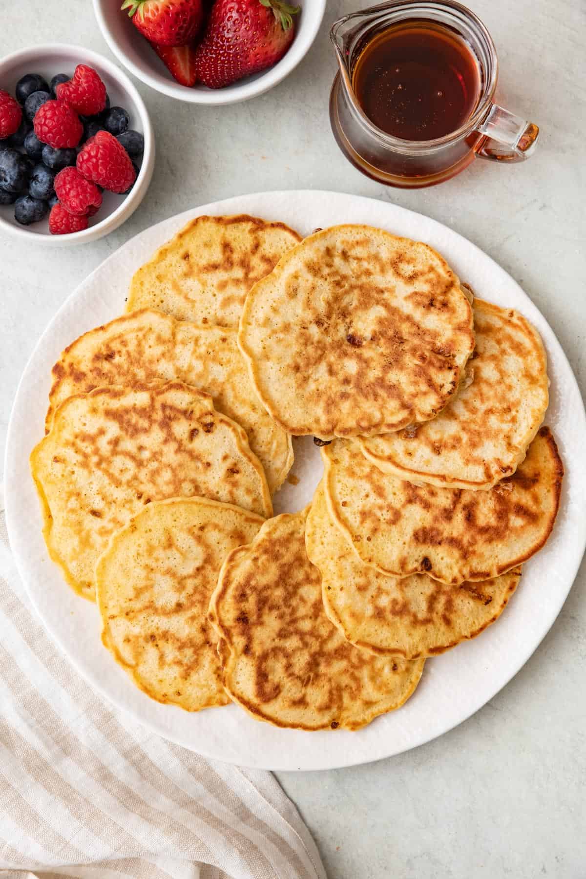 Cottage cheese pancakes layered in a circle overlapping each other on a round plate with two dishes of fresh berries and a side of maple syrup.