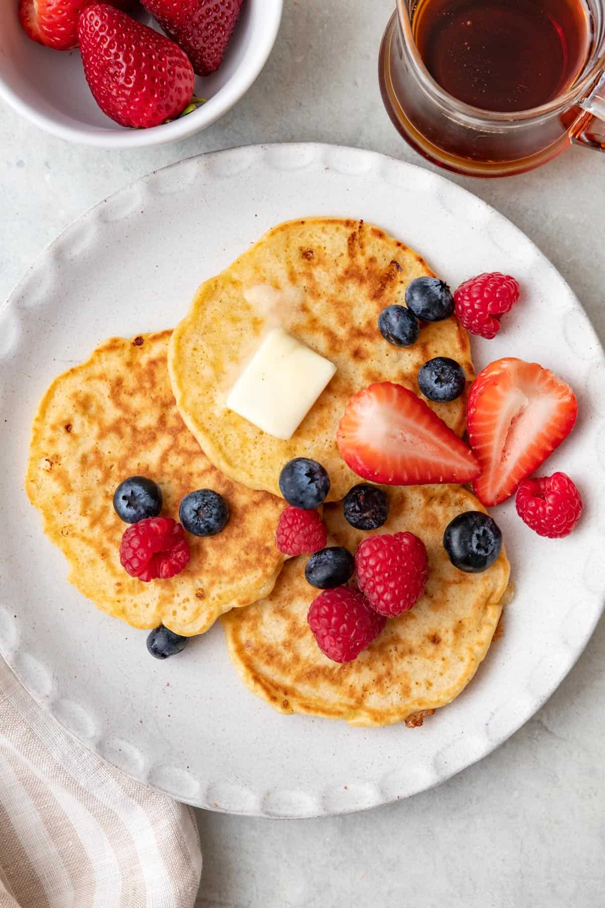 Three cottage cheese pancakes on a plate topped with a pat of butter and fresh berries with maple syrup and more fresh fruit nearby.
