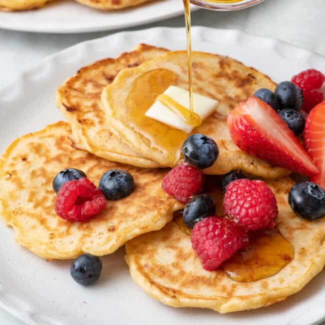 Maple syrup being poured over a plate of cottage cheese pancakes topped with fresh berries and butter with the serving plate of more pancakes nearby.