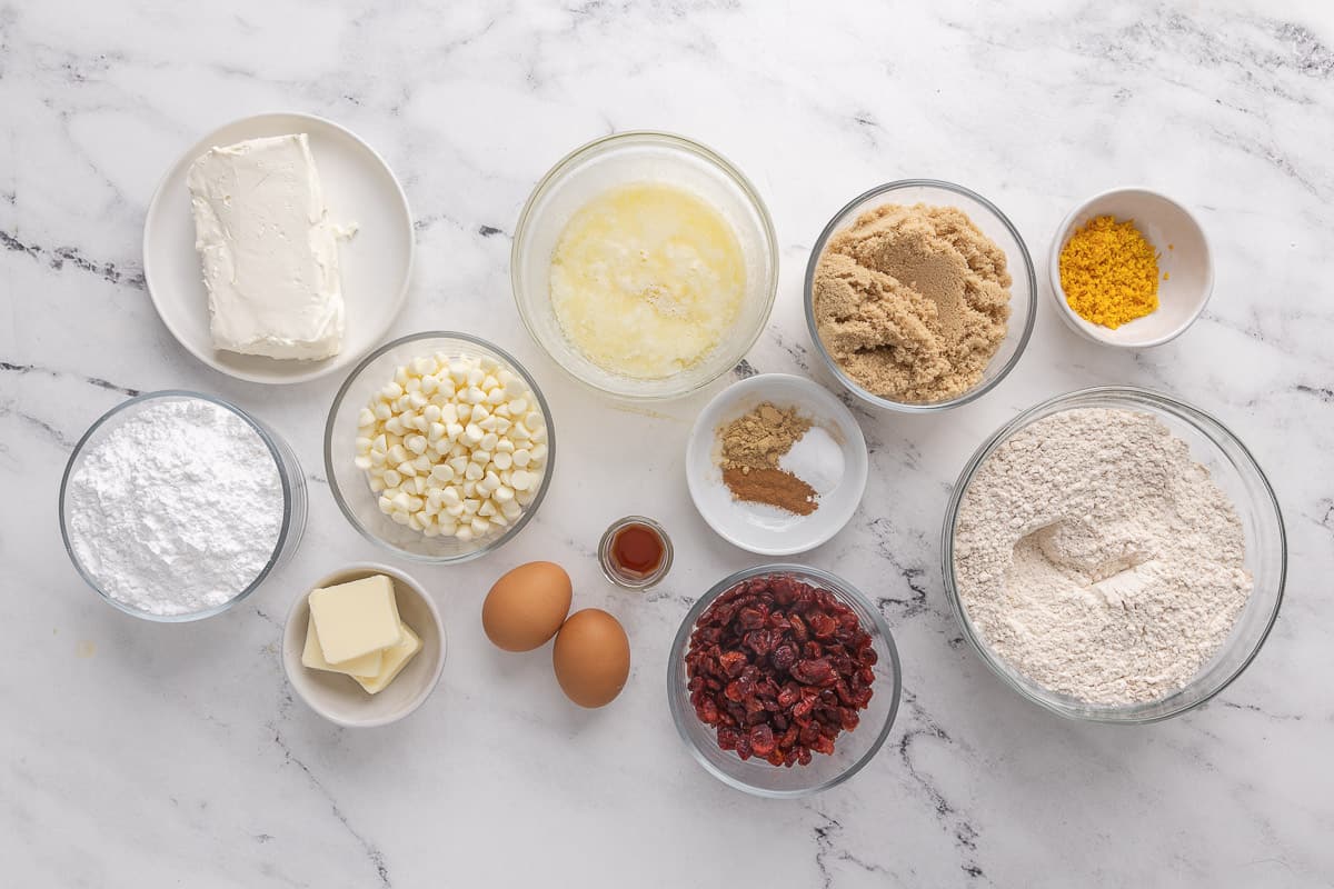 Ingredients for recipe in individual bowls: cream cheese, powdered sugar, white chocolate chips, butter, melted butter, eggs, vanilla, spices, brown sugar, dried cranberries, orange zest, and flour.
