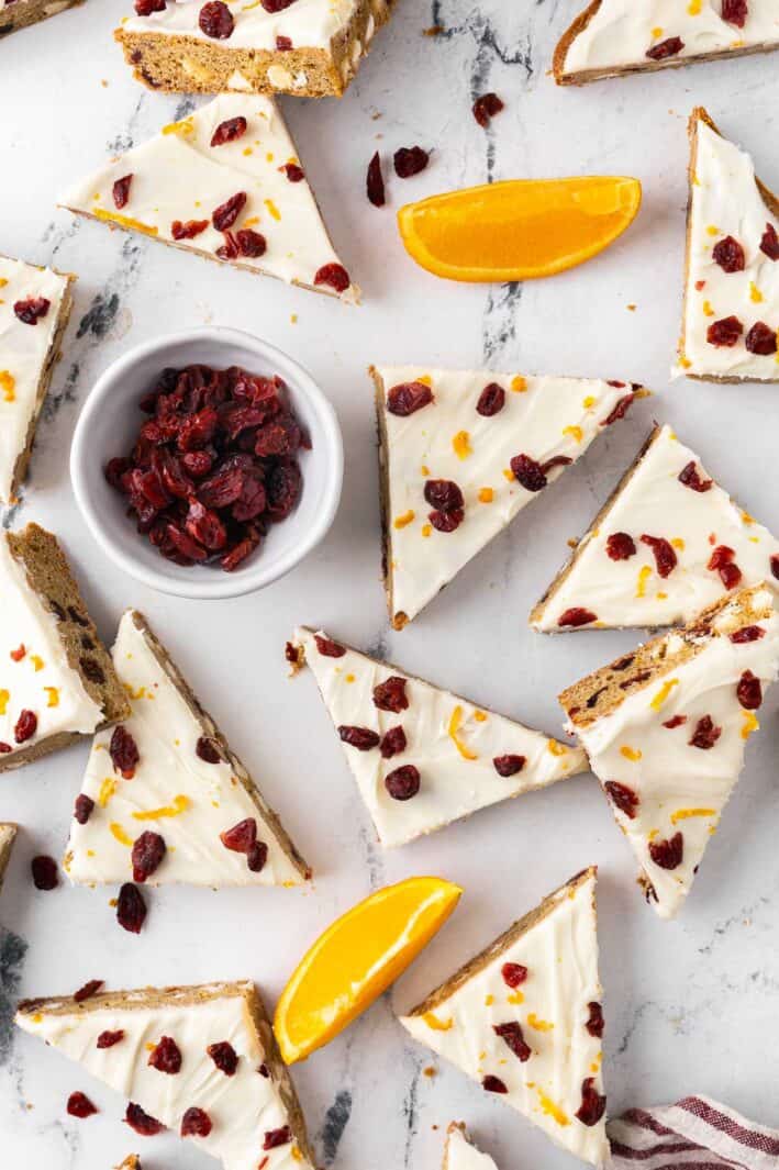 Cranberry bliss bars on a flat surface with a small bowl of dried cranberries and fresh orange slices.