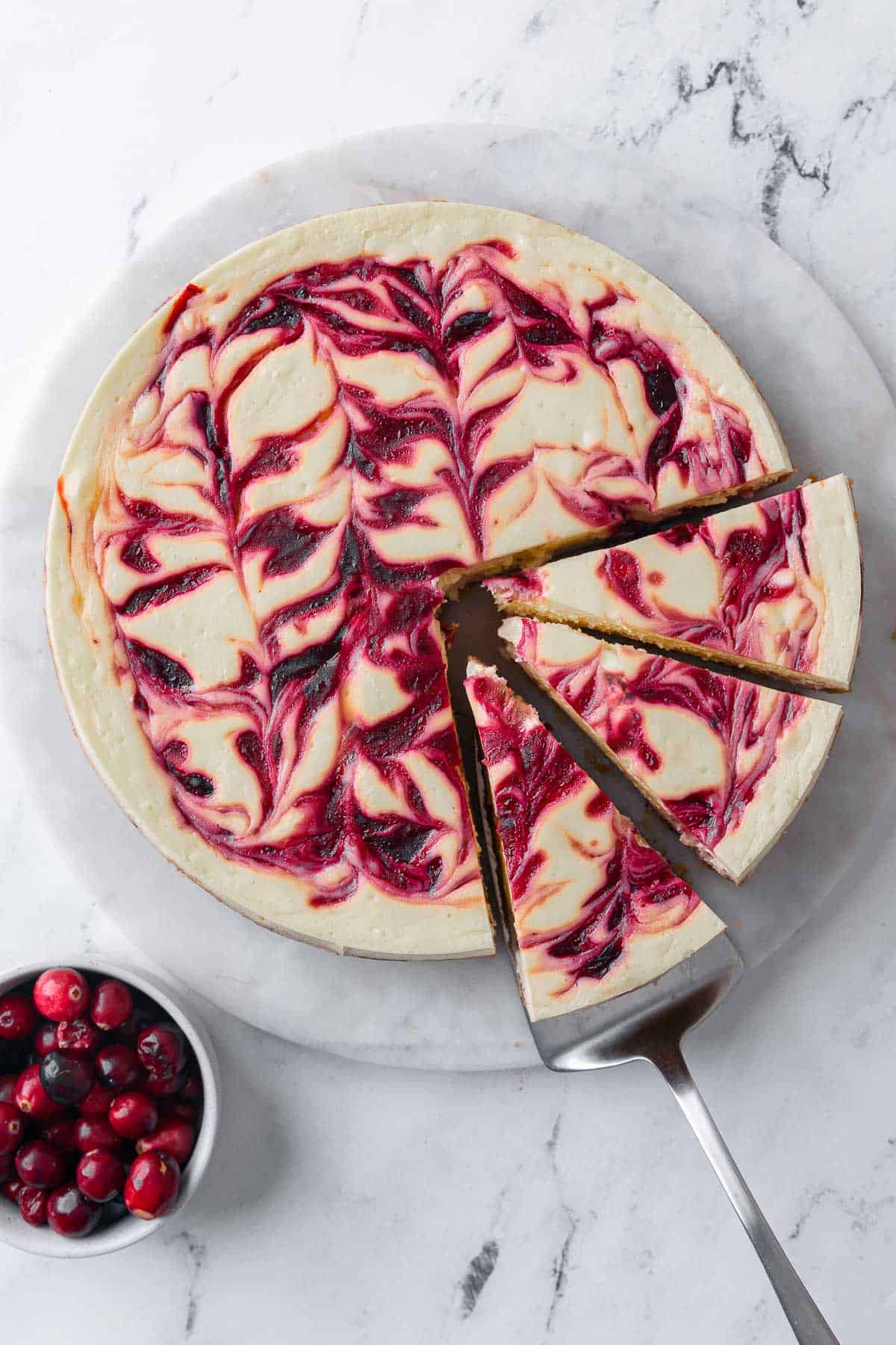 Cranberry cheese cake cut into slices with a serving on a small plate nearby and a small bowl of fresh cranberries nearby.
