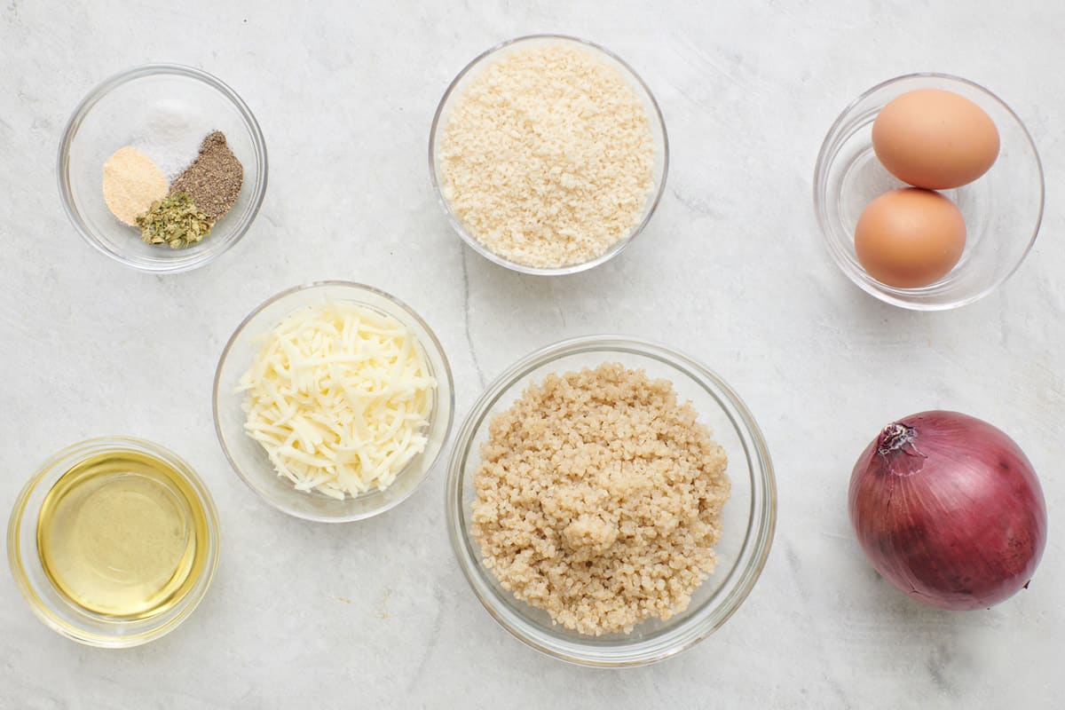Ingredients for recipe in individual bowls: spices, oil, shredded cheese, breadcrumbs, cooked quinoa, 2 eggs, and a red onion.