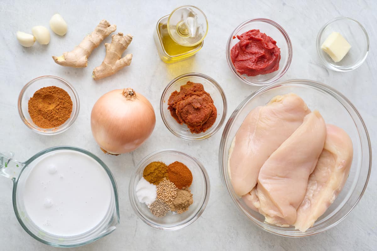 Ingredients for recipe in individual bowls and before prepping: garlic cloves, ginger, curry powder, coconut milk, yellow onion, oil, tomato paste, red curry paste, spice blend, butter, and chicken breasts.