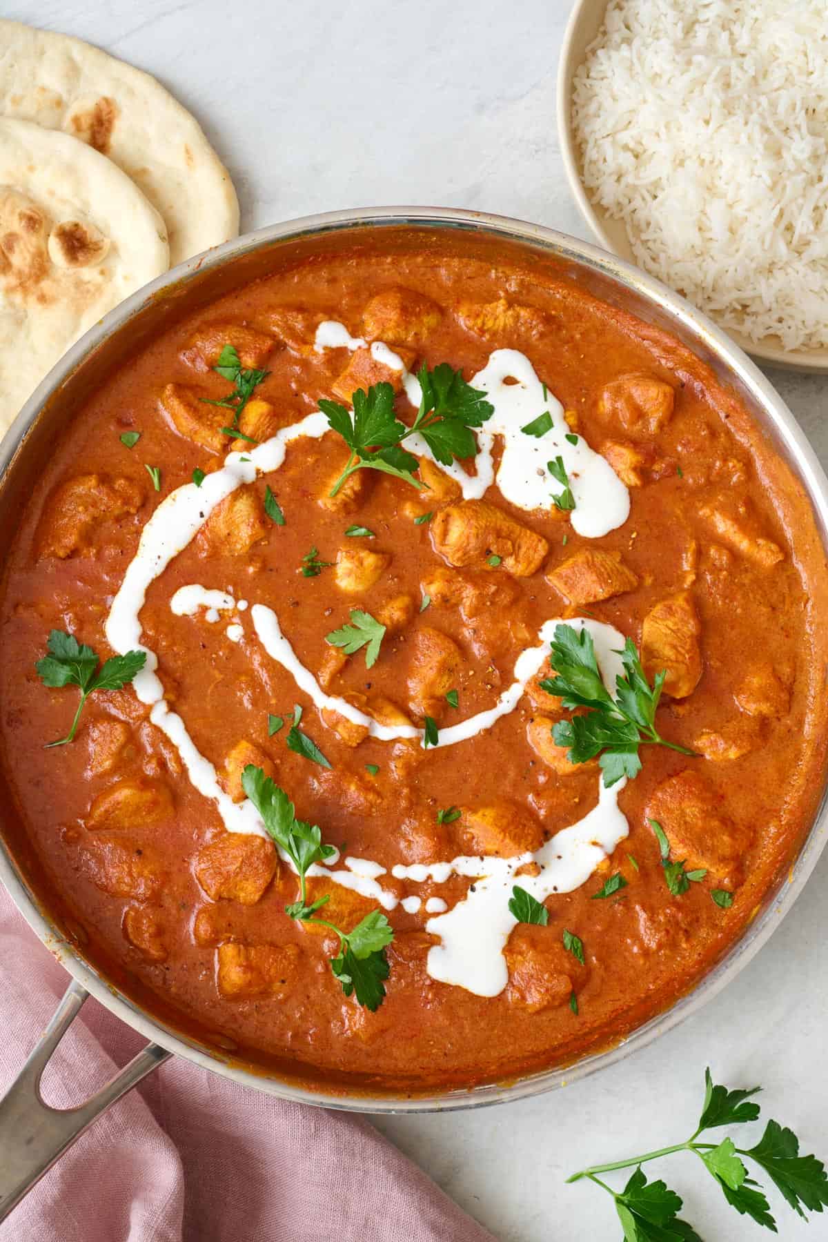 Easy butter chicken in skillet garnished with an extra drizzle of coconut milk, fresh cilantro, and a side of naan and rice nearby.