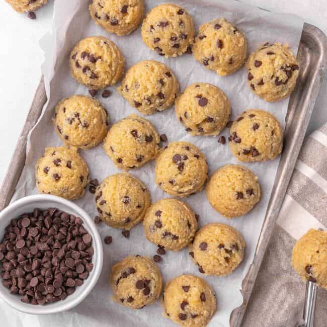 Chocolate chip edible cookie dough balls on a parchment lined sheet pan with a small dish of chocolate chips tucket inside. Edible cookie dough balls in a small high sided white bowl with more around.