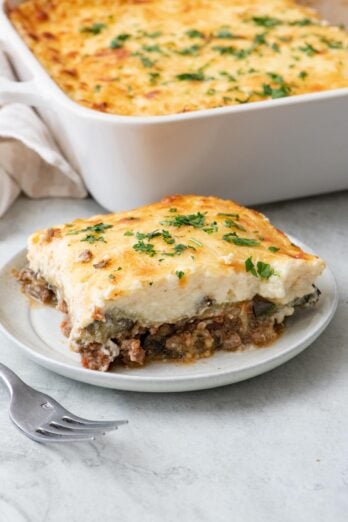 Serving of moussaka on a small plate showing layers of eggplant, beef, and bechamel with a cheesy top next to the baking dish and a fork resting nearby.