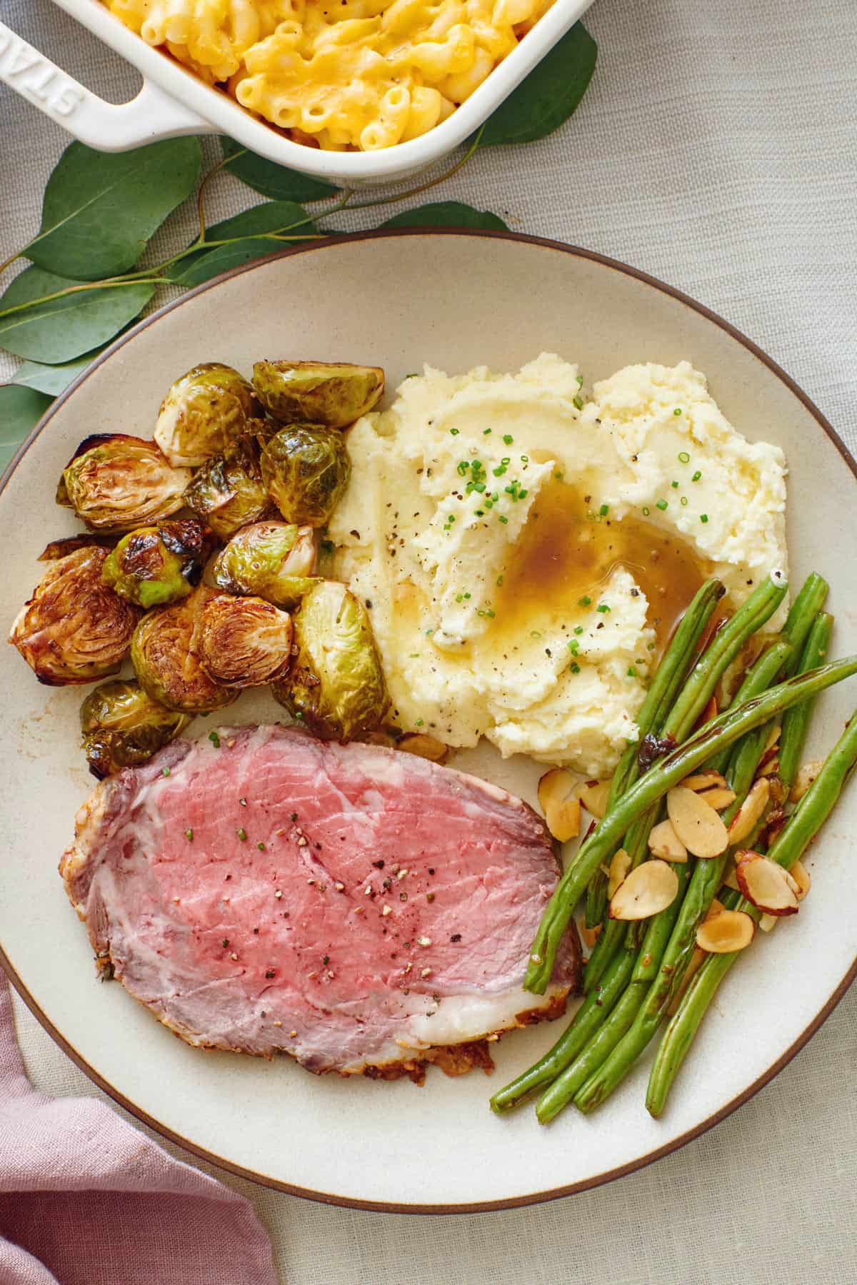 Serving of prime rib on a plate with brussel sprouts, mashed potatoes with au jus, and green beans.