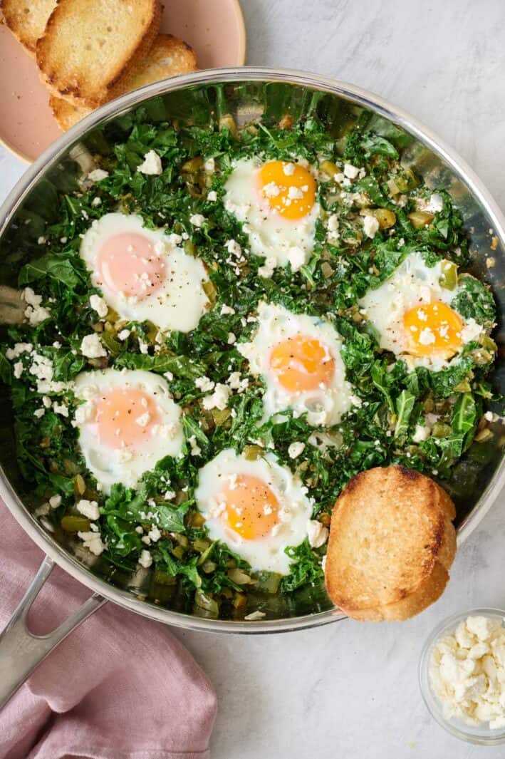 Green Shakshuka made with kale, green peppers and onions, in the skillet with crusty bread on the side and a small bowl of feta nearby.