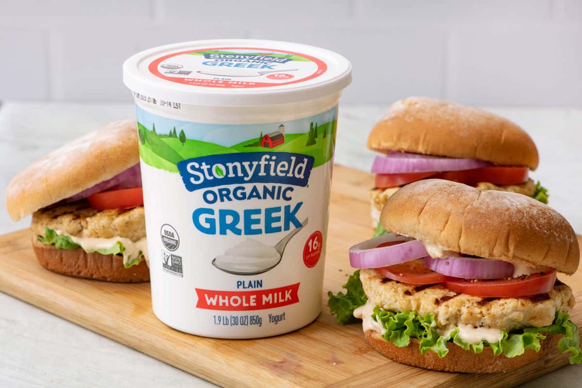 Grilled yogurt chicken burgers assembled with lettuce, tomato, and onion on a cutting board next to a container of Stonyfield organic Greek yogurt.