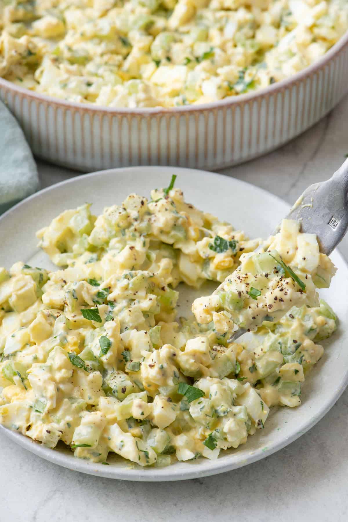 Fork lifting up a bite of egg salad from a small plate with larger serving dish nearby.