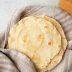 Stack of homemade flour tortillas slightly wrapped in a towel with rolling pin nearby.