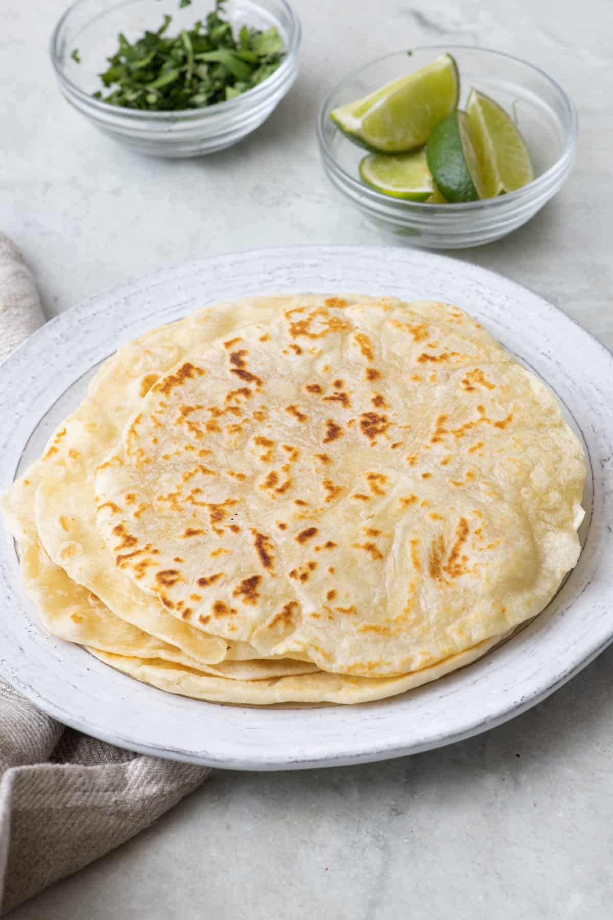 Stack of homemade flour tortillas on a plate with small dishes of lime wedges and chopped cilantro nearby.