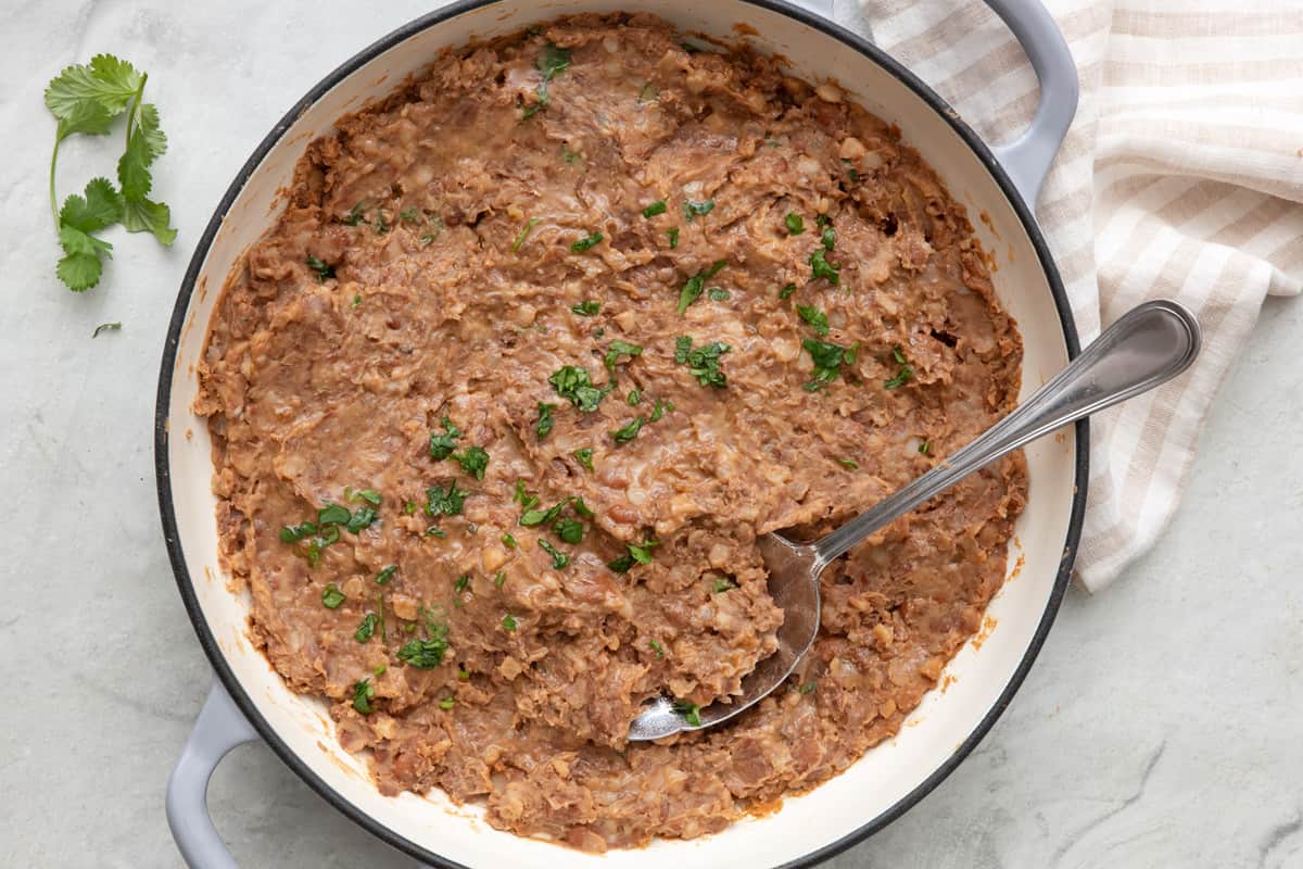 Refried beans in a large pot, garnished with fresh cilantro and a serving spoon dipped in.