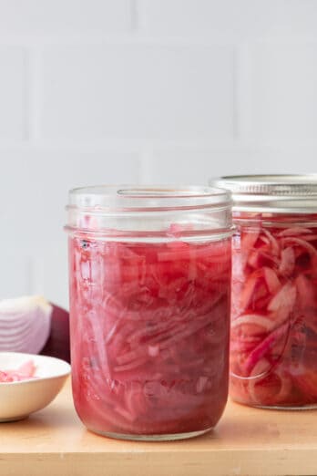 2 jars of pickled red onions, one without a lid and extra red onions nearby.