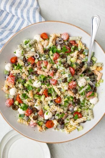 Italian pasta salad in a large serving bowl with a spoon dipped in a serving plates nearby.